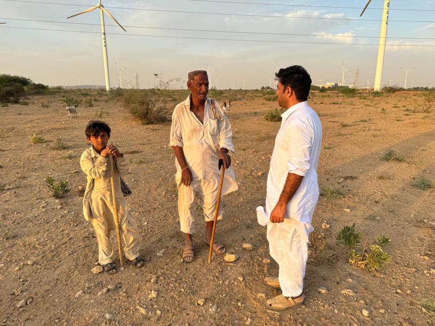 Three people—an elderly man, a young boy, and another adult man—stand in conversation on a dry, open landscape scattered with low shrubs. They wear traditional light-colored clothing and hold wooden walking sticks. In the background, multiple large wind turbines rise across the horizon under a partly cloudy sky, suggesting a juxtaposition of local community life and renewable energy development. A few goats can be seen grazing in the distance.