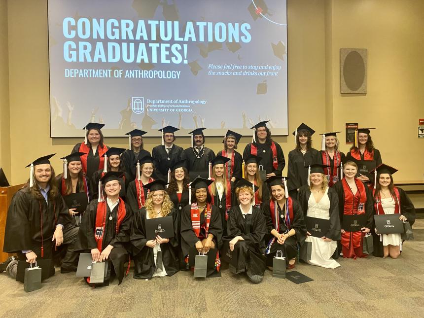 Alt text: A group of University of Georgia Anthropology graduates pose together in their caps and gowns in front of a large screen that reads “Congratulations Graduates! Department of Anthropology.” The group includes about two dozen smiling students, some holding diploma covers and gift bags, standing and kneeling in rows in a classroom or lecture hall.