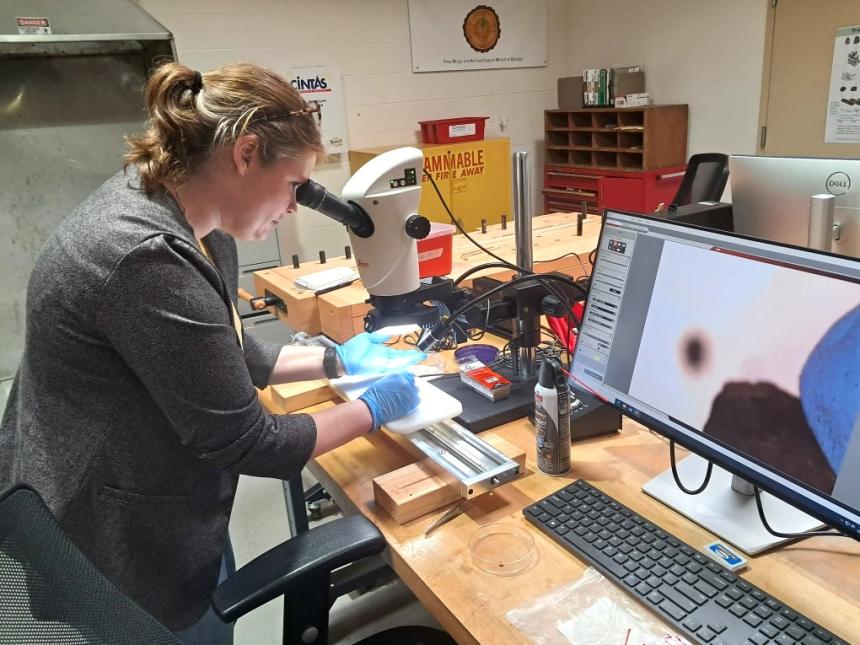Dr. Brita Lorentzen wearing coat and gloves prepares a wood sample under a microscope in a laboratory. A nearby computer monitor displays a magnified image of the sample. The workspace includes lab tools, a keyboard, and safety equipment, with cabinets and lab signage in the background, including a flammable materials warning and a Tree Ring and Archaeological Wood of Georgia banner