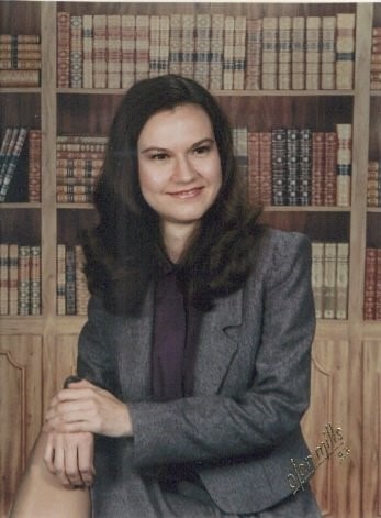 A person in a suit sitting in front of a bookshelf</p>
<p>Description automatically generated