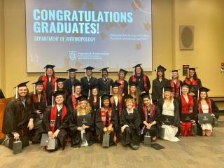 Alt text: A group of University of Georgia Anthropology graduates pose together in their caps and gowns in front of a large screen that reads “Congratulations Graduates! Department of Anthropology.” The group includes about two dozen smiling students, some holding diploma covers and gift bags, standing and kneeling in rows in a classroom or lecture hall.