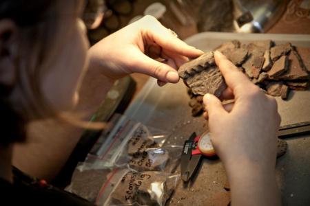 A person examines a decorated pottery sherd at an archaeological lab table. The table is covered with various pottery fragments, some labeled in plastic bags, a caliper for measuring, and other lab tools used for artifact analysis