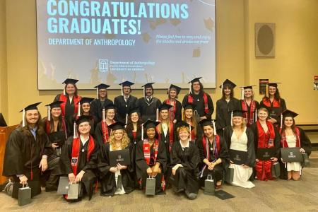 Alt text: A group of University of Georgia Anthropology graduates pose together in their caps and gowns in front of a large screen that reads “Congratulations Graduates! Department of Anthropology.” The group includes about two dozen smiling students, some holding diploma covers and gift bags, standing and kneeling in rows in a classroom or lecture hall.
